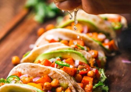 The image shows a hand squeezing lime over a row of tacos filled with vegetables and chickpeas, placed on a wooden board with lime slices.