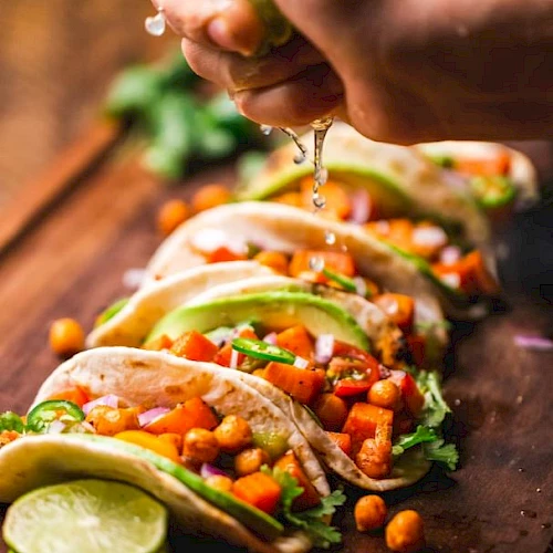 The image shows a hand squeezing lime over a row of tacos filled with vegetables and chickpeas, placed on a wooden board with lime slices.