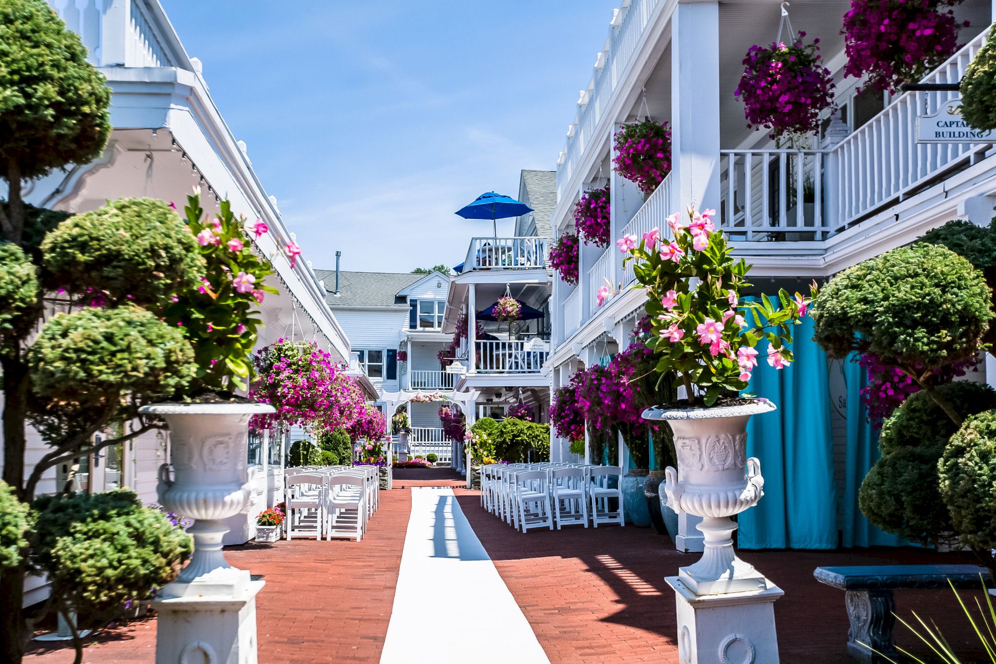 A picturesque courtyard with white buildings, flower pots, hanging baskets, trimmed bushes, and outdoor seating under a clear blue sky.