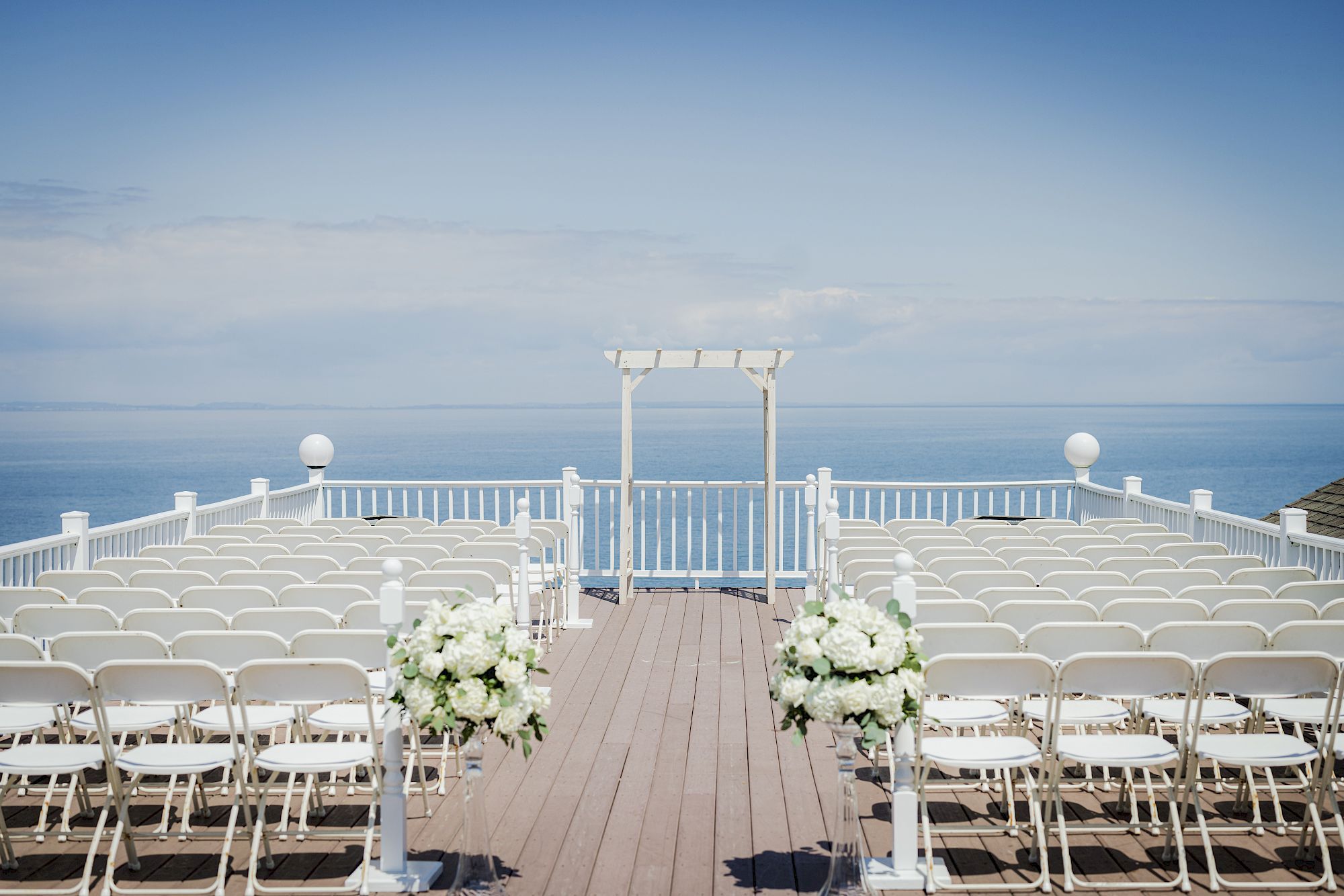 An outdoor wedding setup with white chairs arranged in rows, floral decorations, and a wooden arch with an ocean view in the background.