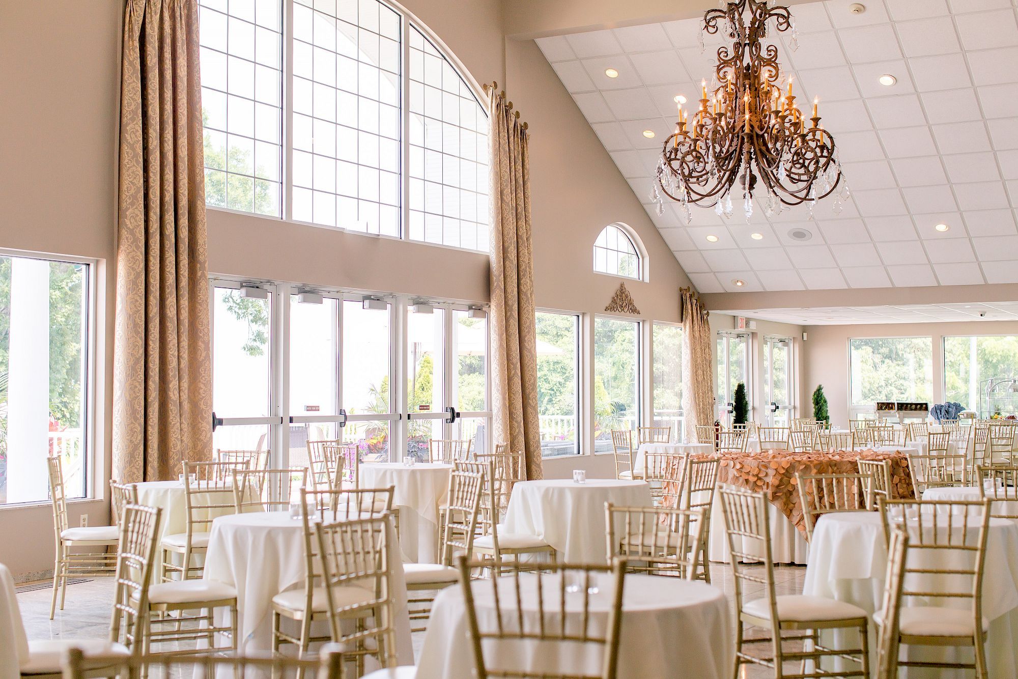 A brightly lit banquet hall with large windows, round tables with white tablecloths, and elegant gold chairs under a decorative chandelier.