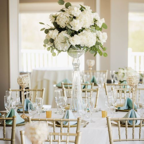 The image shows an elegantly set dining table with a tall centerpiece of white flowers, gold chairs, and glasses neatly arranged around the table.