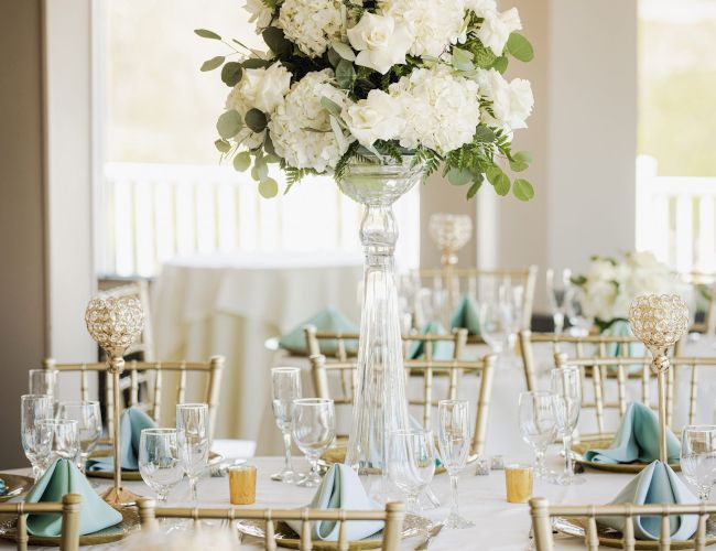 The image shows an elegantly set dining table with a tall centerpiece of white flowers, gold chairs, and glasses neatly arranged around the table.