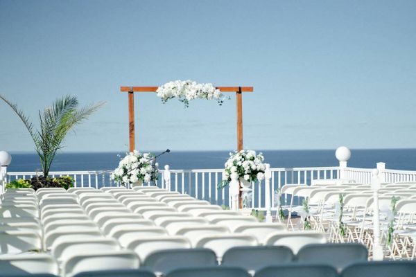 An outdoor wedding setup with rows of white chairs facing a wooden arch adorned with white flowers and the ocean in the background.