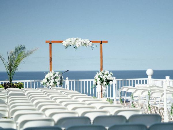 An outdoor wedding setup with rows of white chairs facing a wooden arch adorned with white flowers and the ocean in the background.