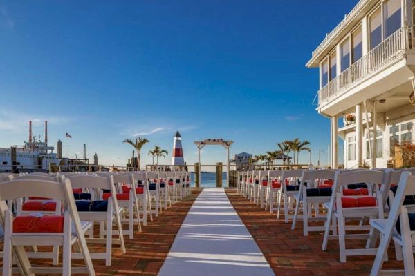 The image shows an outdoor wedding setup with white chairs lined up, an aisle runner, and a view of a lighthouse, boats, and a building on the right.