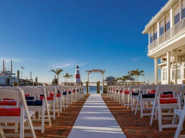 The image shows an outdoor wedding setup with white chairs lined up, an aisle runner, and a view of a lighthouse, boats, and a building on the right.