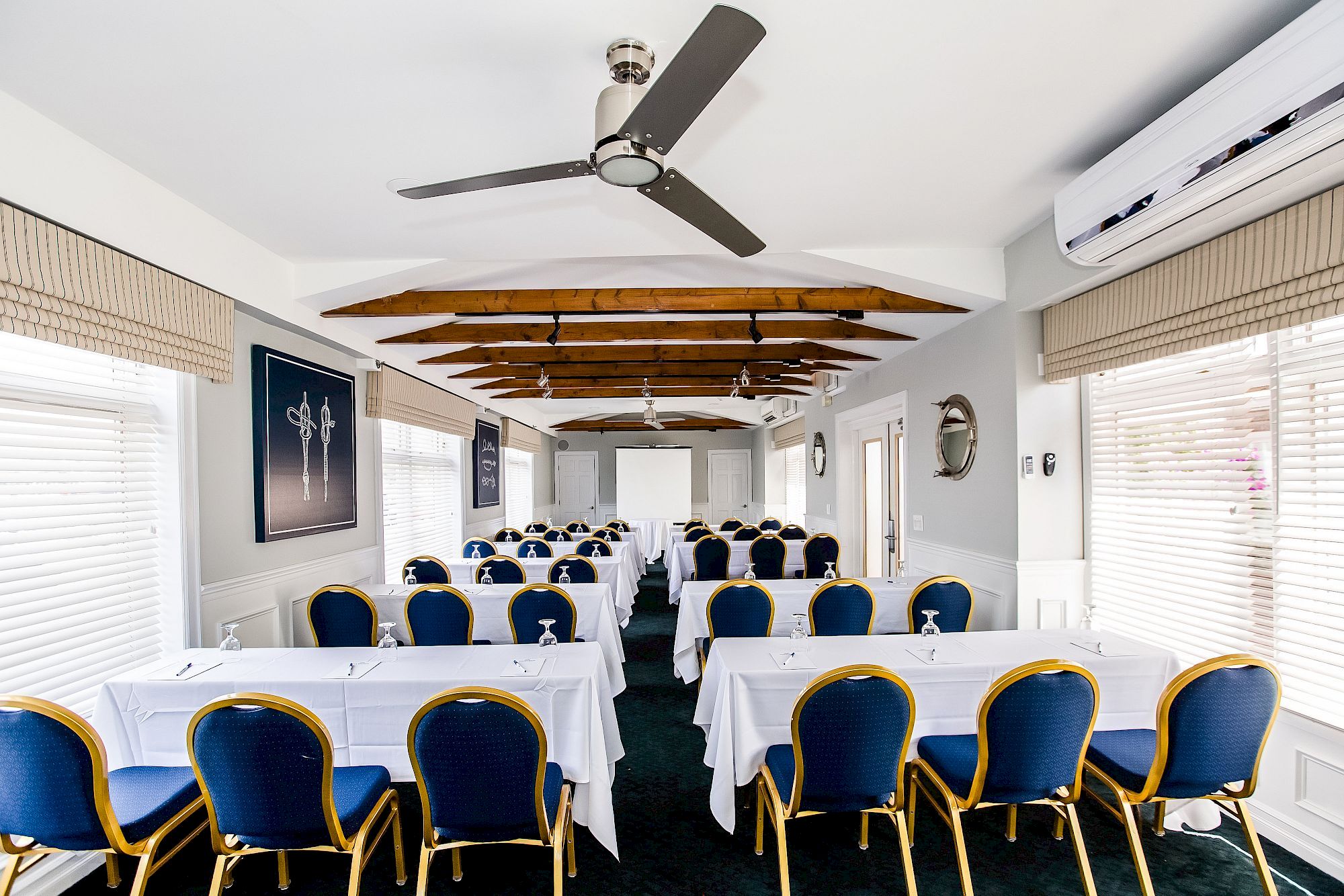 A conference room with arranged tables and blue chairs, a ceiling with wooden beams, and large windows on both sides covered with blinds.