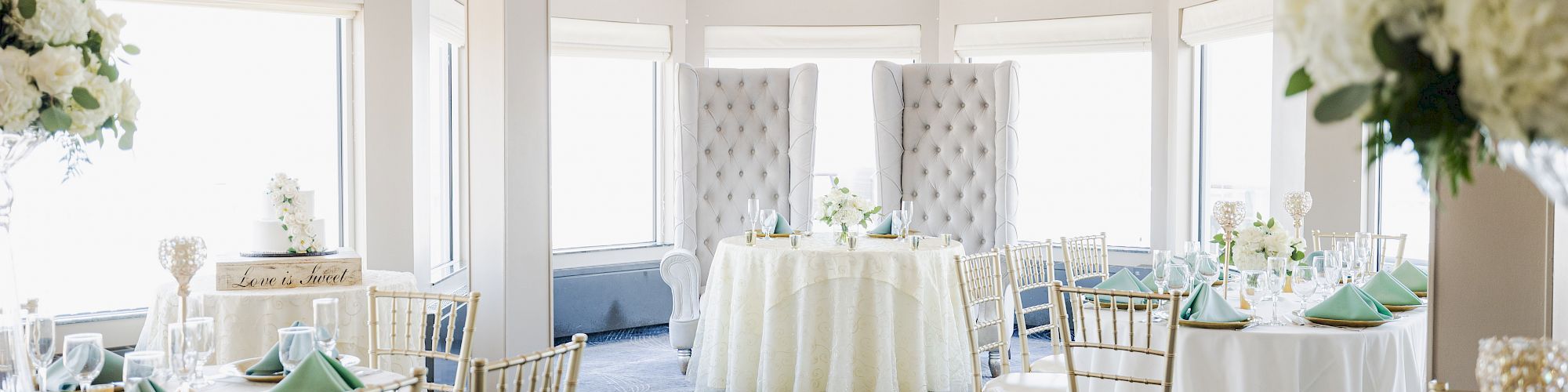 A beautifully decorated banquet hall with elegant dining tables, green napkins, floral arrangements, and large windows is shown.