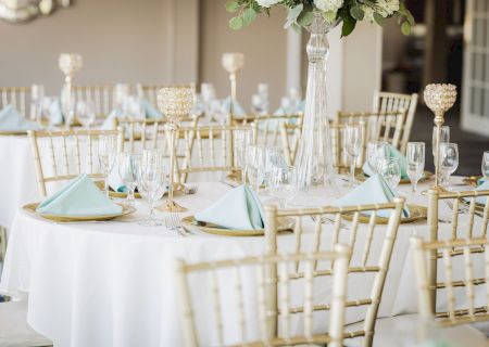 An elegant table setting with white tablecloths, gold chairs, teal napkins, glassware, and floral centerpieces in crystal vases.