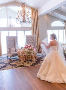 A bride in a wedding gown stands in a decorated reception hall with chandeliers, floral arrangements, and elegant seating, preparing for the event.