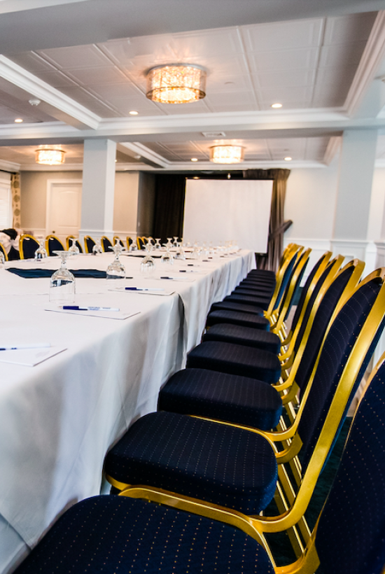 A conference room set up with long tables, blue and gold chairs, white tablecloths, glassware, notepads, pens, and a projector screen in the background.