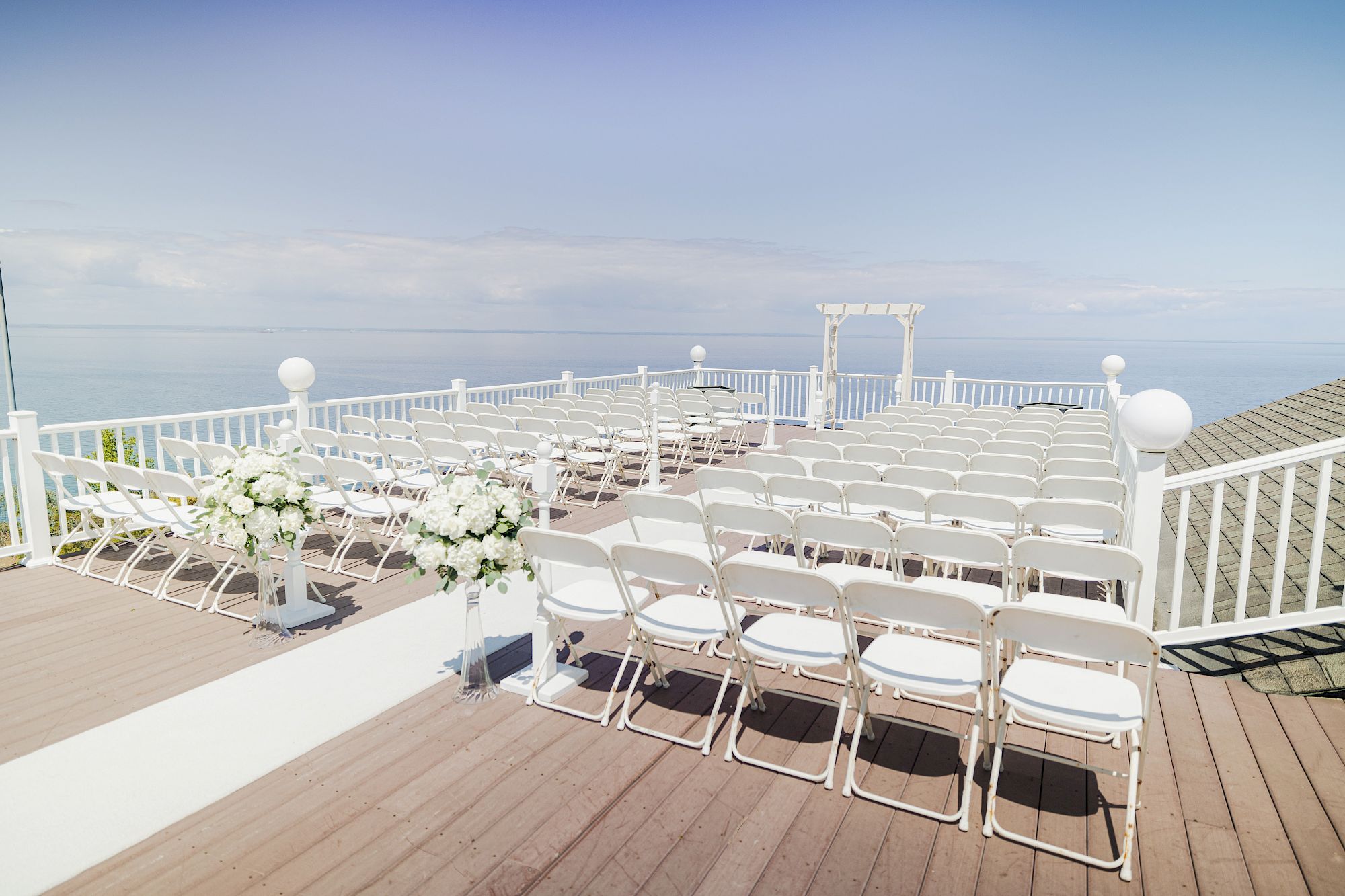 An outdoor wedding setup overlooking the water, with white chairs arranged in rows, floral decorations, and an archway at the front, on a wooden deck.