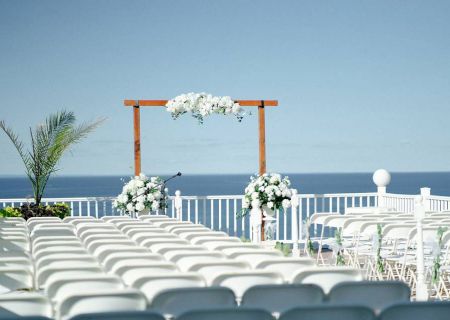 The image shows an outdoor wedding setup with white chairs arranged in rows, a floral arch, and a backdrop of the ocean.