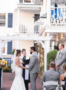 A couple is getting married outdoors, with bridesmaids, groomsmen, and guests seated around them. White buildings are visible in the background.