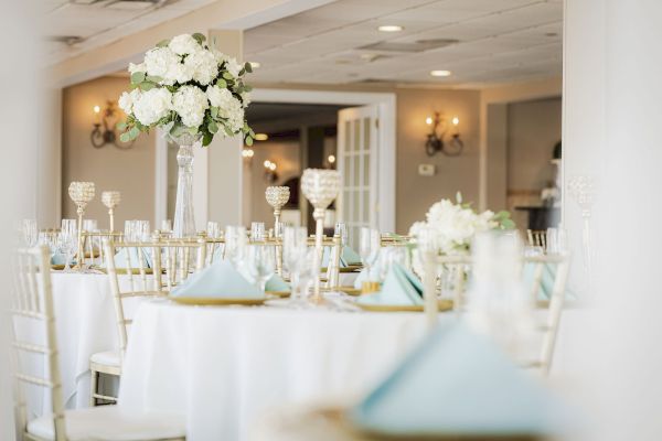 This image shows an elegantly set banquet hall with white tablecloths, floral centerpieces, light blue napkins, and crystal decor on the tables.