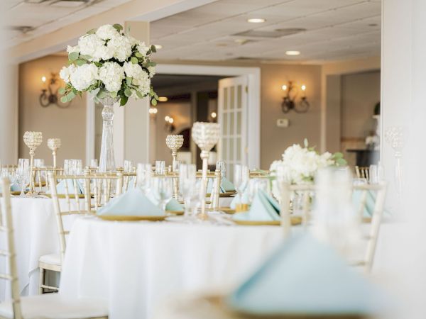 This image shows an elegantly set banquet hall with white tablecloths, floral centerpieces, light blue napkins, and crystal decor on the tables.