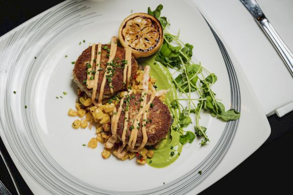 The image shows a plated dish with two crab cakes topped with sauce, garnished with greens, corn, and a grilled lemon half on a patterned plate.