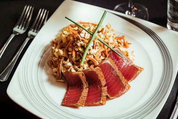 A plate with sliced seared tuna and a mixed salad garnished with chives. Forks are placed beside the plate, and a glass is visible in the background.