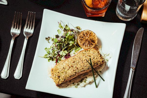 A plated meal consists of what appears to be a breaded fish fillet, garnished with a lemon slice, chives, and a side salad.