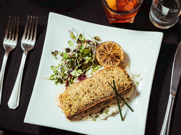 A plated meal consists of what appears to be a breaded fish fillet, garnished with a lemon slice, chives, and a side salad.