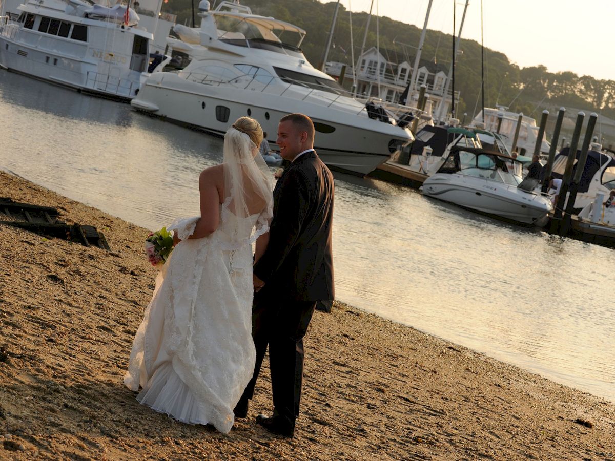 A couple in wedding attire stands on a sandy shore, facing a harbor with yachts and boats docked in the background.