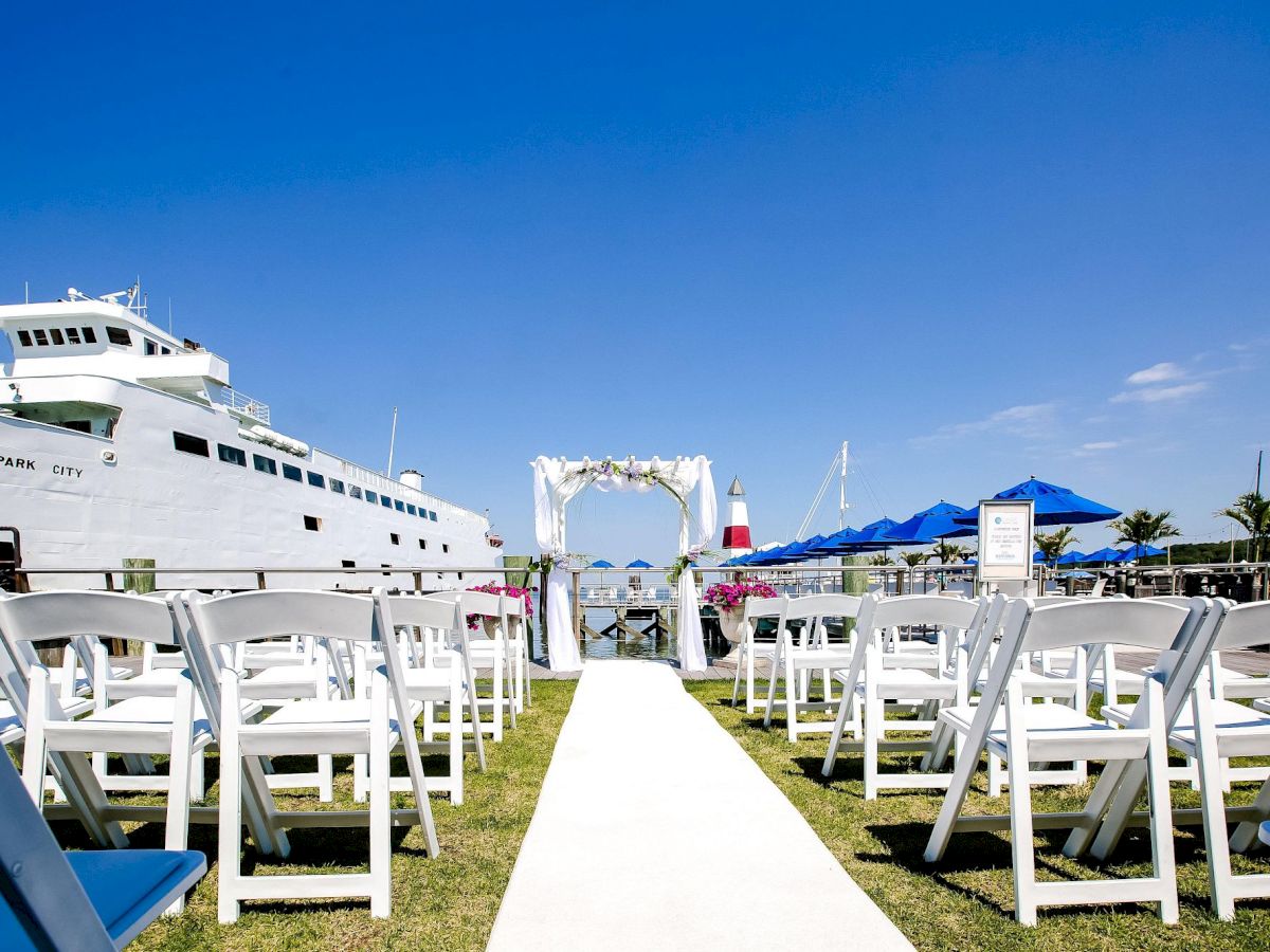 An outdoor wedding setup with white chairs arranged near a large boat, under a clear blue sky, featuring a white archway in the distance.