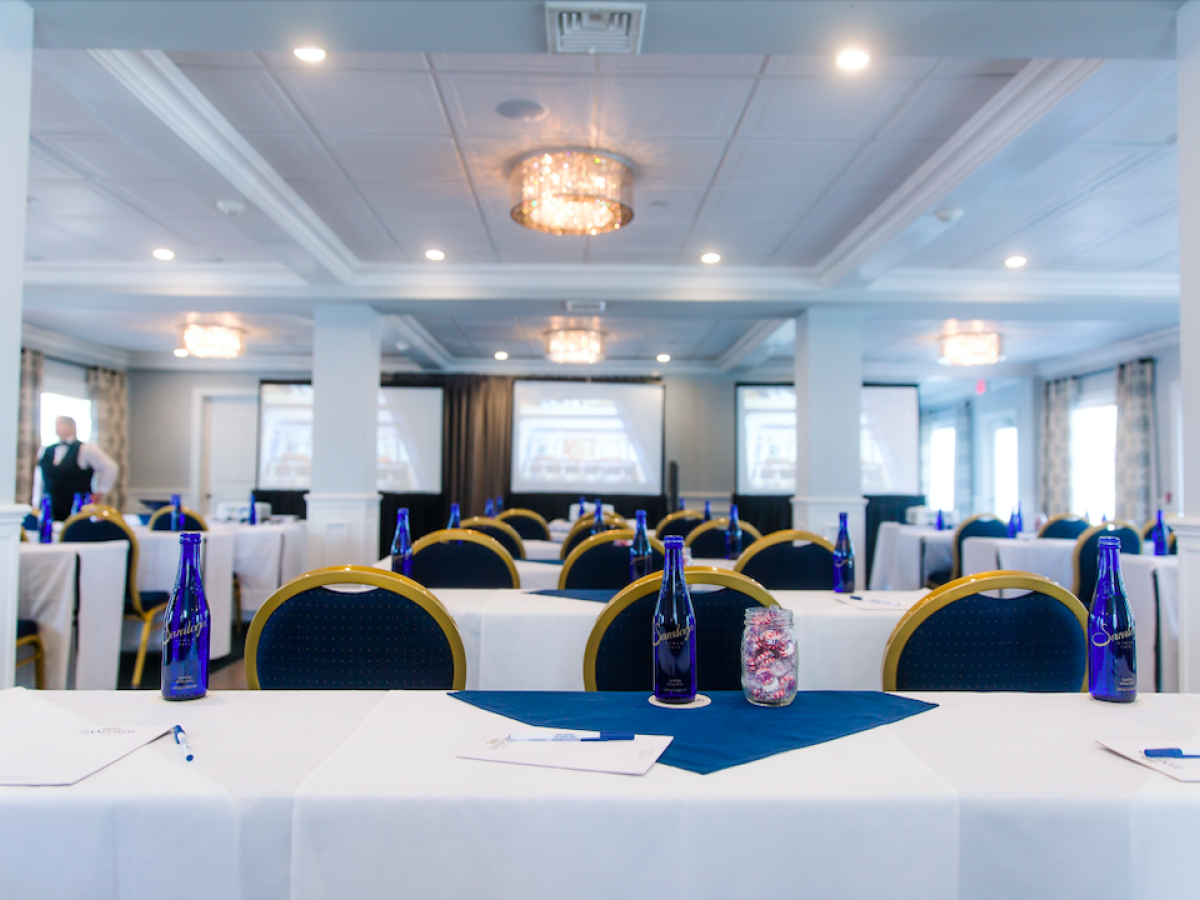 A conference room with rows of tables and chairs, blue water bottles, notebooks, pens, and screens projected at the front is shown.