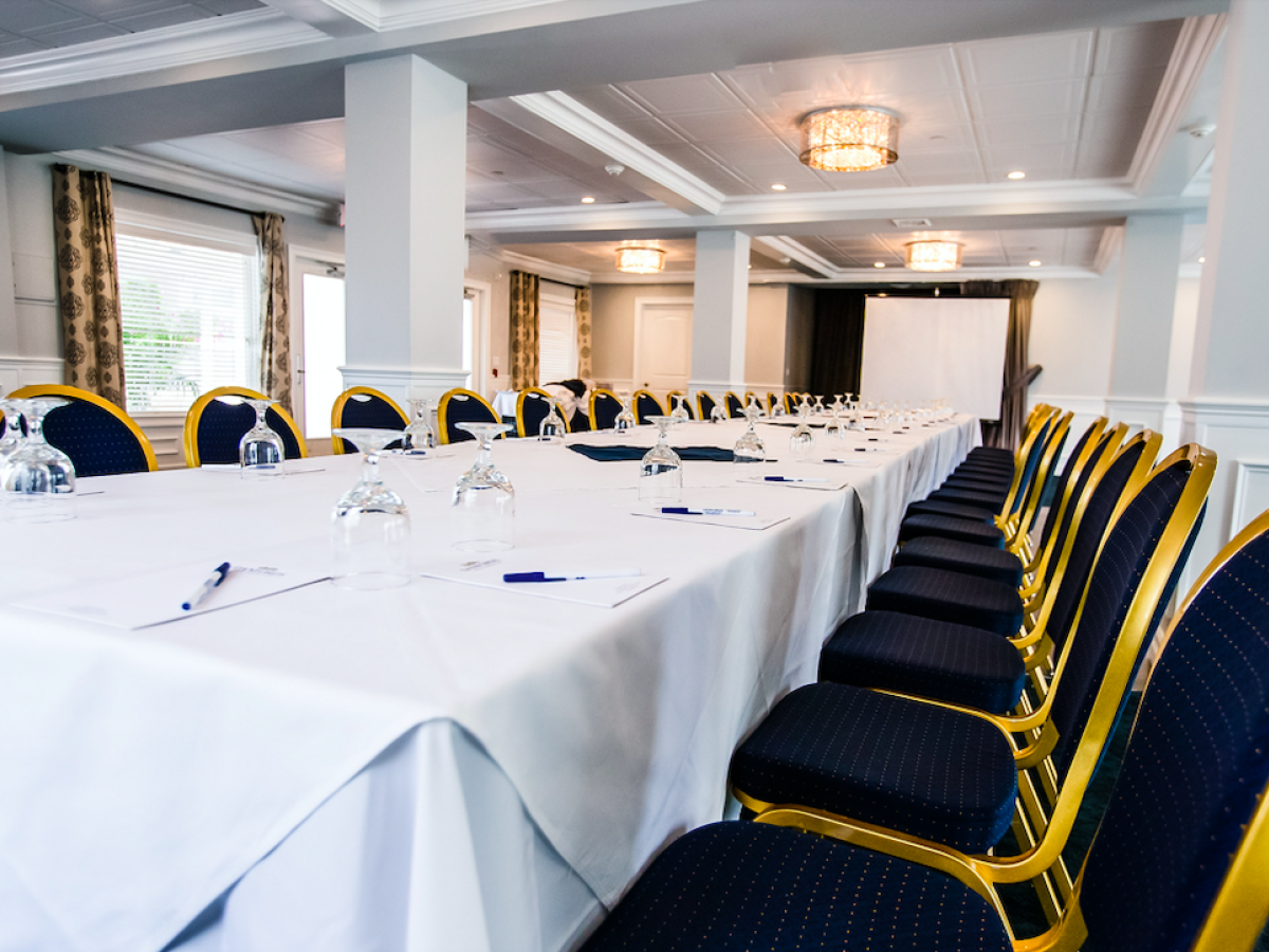 The image shows a conference room with a long table covered in white cloth, surrounded by blue chairs, and set with pens, papers, and water glasses.