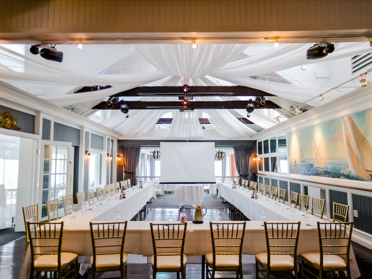 A well-decorated conference room with a U-shaped table setup, elegant chairs, white drapes, and a large projector screen at the front ending the sentence.