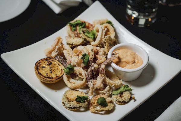 A plate of fried calamari garnished with herbs, served with a grilled lemon half and a small bowl of dipping sauce on the side.