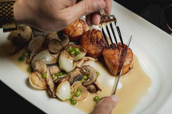 A person cutting into seared scallops on a plate with sautéed mushrooms, onions, peas, and sauce.