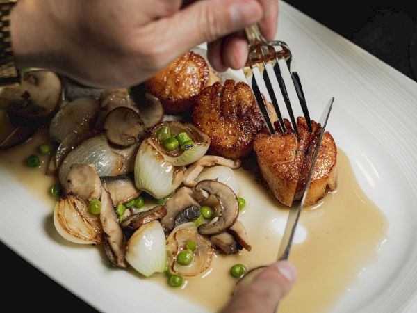 A person cutting into seared scallops on a plate with sautéed mushrooms, onions, peas, and sauce.