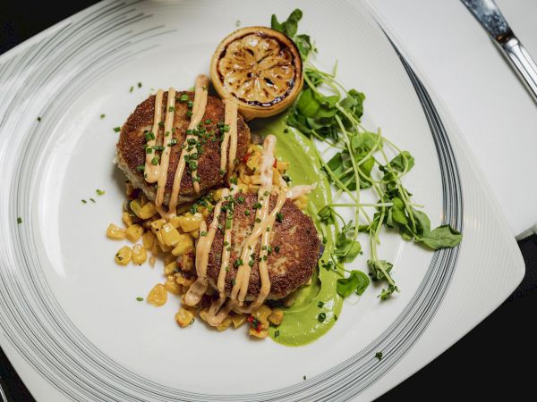 This image shows a plate with two crab cakes, garnished with sauce, grilled lemon, microgreens, and corn relish, served on a decorative white plate.