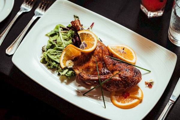 A roasted chicken garnished with orange slices, herbs, and accompanied by a side salad on a white plate.