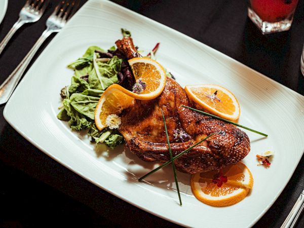 A roasted chicken garnished with orange slices, herbs, and accompanied by a side salad on a white plate.