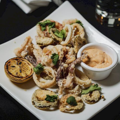 The image shows a plate of fried calamari served with a lemon half and a small bowl of dipping sauce, garnished with cilantro.