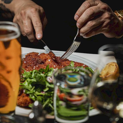 A person is cutting into a breaded dish, possibly schnitzel, on a plate with salad and bread, with a beverage and wine glass nearby, hands visible.