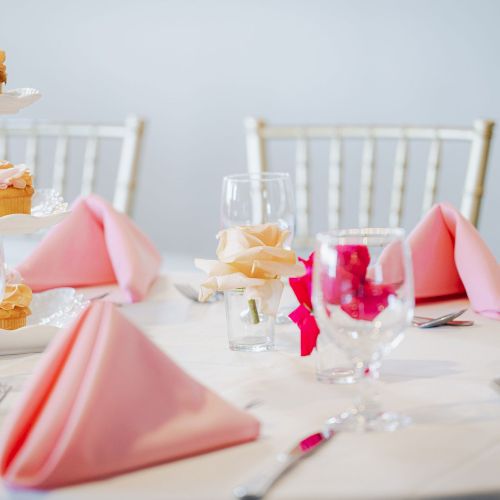 A table set for a formal event with pink napkins, glassware, silverware, and a tiered tray with cupcakes. There are gold-backed chairs around the table.