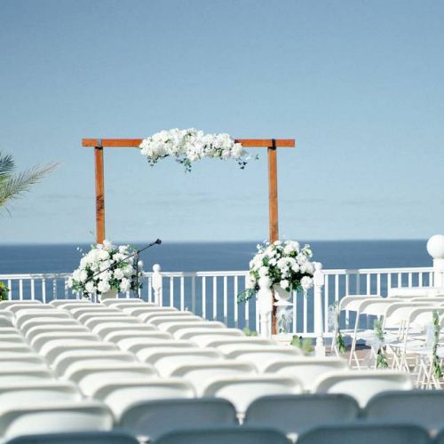 Rows of white chairs face an arch decorated with flowers, set up for an outdoor event overlooking the ocean.