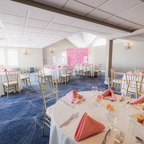 The image shows a decorated event space with round tables covered in white tablecloths, pink napkins, and cupcakes on stands.