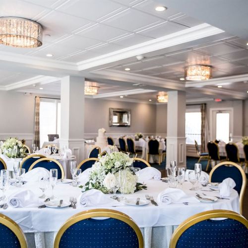 A banquet hall with round tables set for dining, featuring white linens, floral centerpieces, and blue chairs. Chandeliers hang from the ceiling.