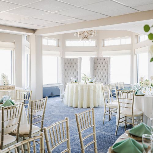 Elegant banquet hall setup with gold chairs, round tables adorned with white linens, green napkins, and floral centerpieces, bathed in natural light.