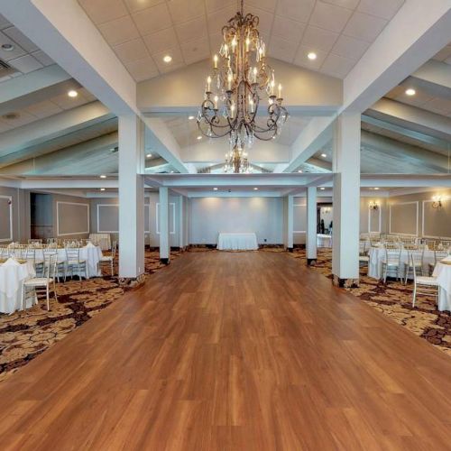 A spacious banquet hall with elegant chandeliers, white draped tables, and a wooden dance floor, ready for an event or celebration.