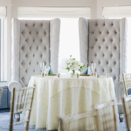 A round table with a white tablecloth is set with glassware and a floral centerpiece, flanked by two tall, tufted chairs near large windows.