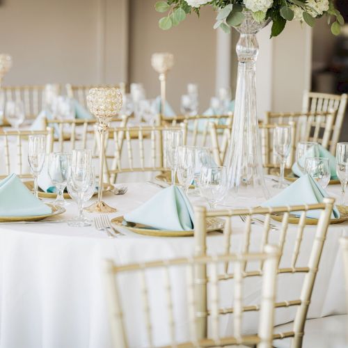 The image depicts a beautifully set dining table with white tablecloths, gold chairs, blue napkins, and elegant floral centerpieces.