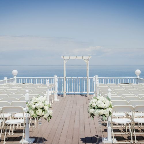 An outdoor wedding setup on a deck facing the sea with white chairs, floral arrangements, and a white arch at the end of the aisle.