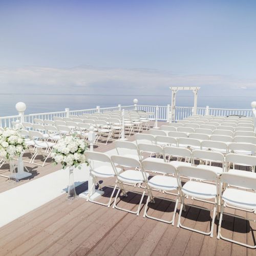 An outdoor wedding ceremony setup on a deck overlooking the ocean, featuring white chairs arranged in rows and a floral arrangement.