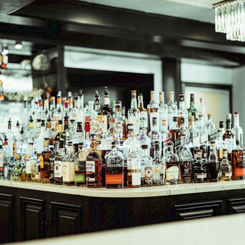 The image shows a bar counter with a large assortment of different liquor bottles arranged on top of it, in a well-lit and modern setting.