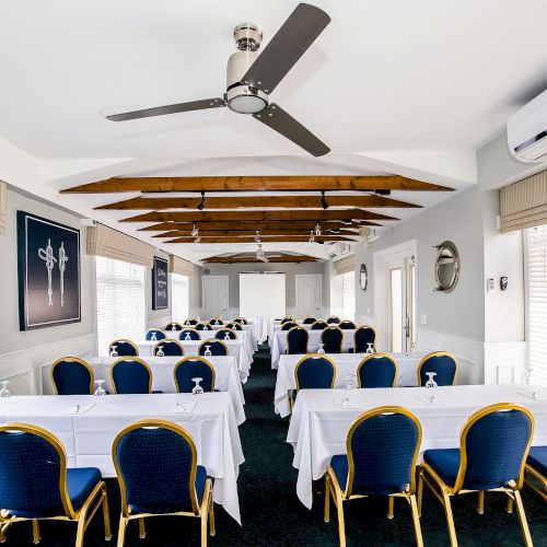 The image shows a conference room setup with blue and gold chairs, white tablecloths, ceiling fans, and a projector screen at the front.